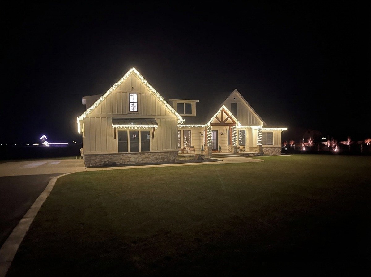 White farmhouse in Rexburg, Idaho with warm white lights on the peaks, eaves and porch columns at night