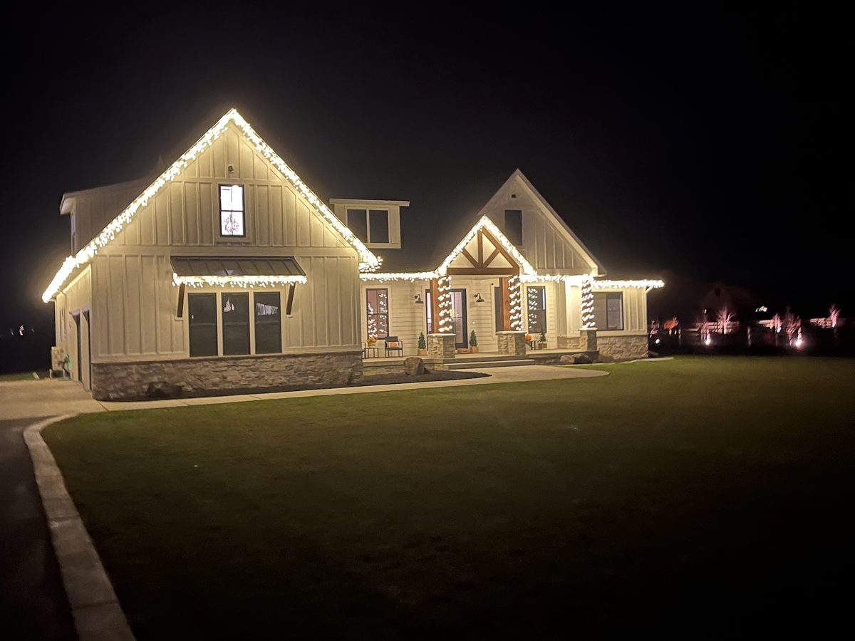 White farmhouse in Rexburg, Idaho with warm white lights on the peaks, eaves, side eave and porch columns at night