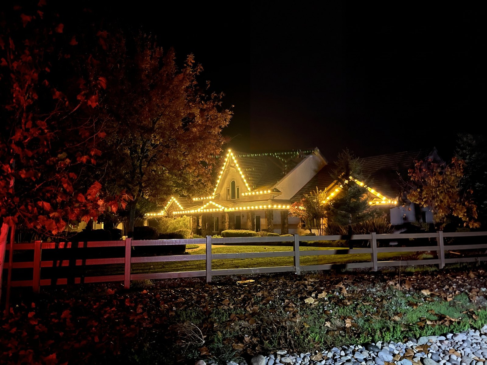 Wilton home with warm white C9 lights on every gable and roofline at night, seen across a rail fence