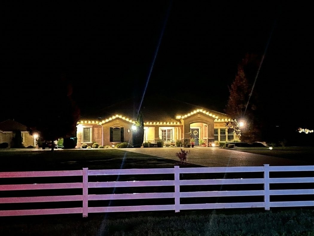 Home behind a white rail fence with C9 lights on the roofline at night
