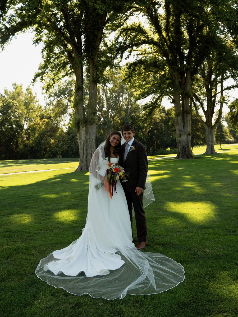 Zach and Brooklyn on their wedding day, standing on the grass under tall shade trees, her long veil spread out behind her
